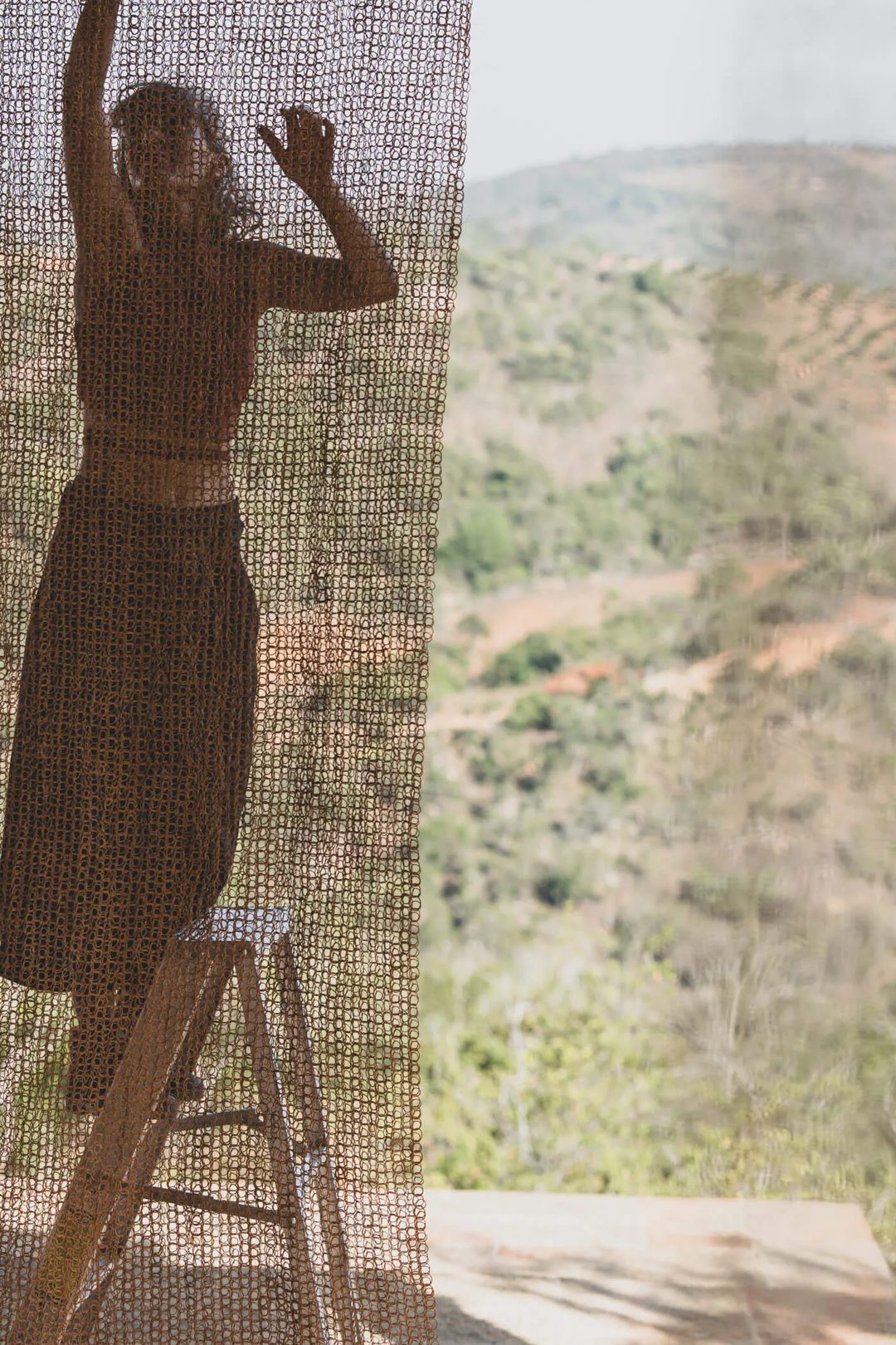 Silhouette of a woman on a ladder hanging Nodomondo sunshade netting with a rural landscape visible in the background.
