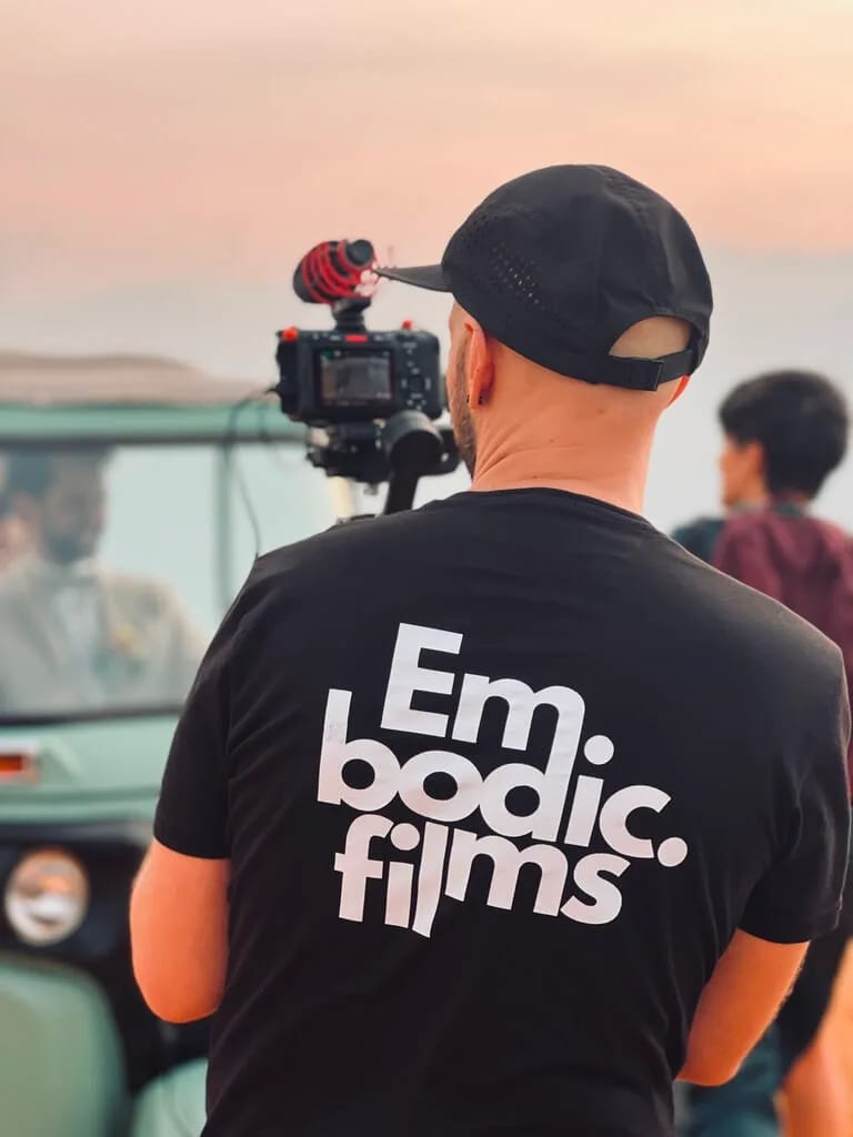 A person wearing a black cap and a black "Embodic Films" shirt operates a camera mounted on a tripod, with a blurred car and other individuals in the background.