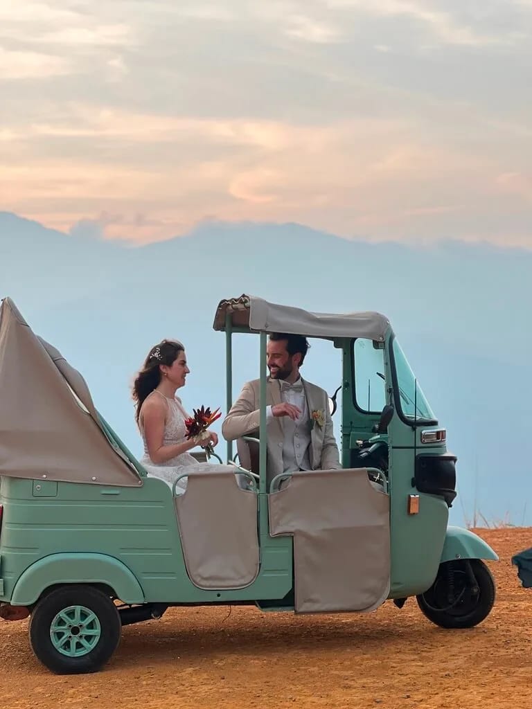 A couple in wedding attire sits in a vintage green three-wheeled vehicle on a dirt road with mountains and a cloudy sky in the background. The bride holds a small bouquet.