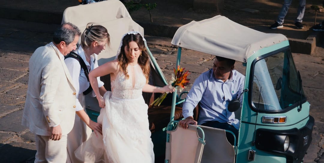 A bride in a white dress steps out of a tuk-tuk, assisted by two men and followed by another woman, holding a bouquet of orange and yellow flowers.