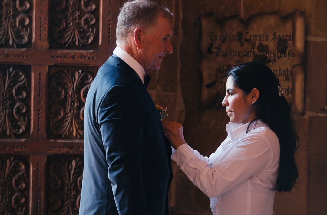 A person in a white shirt is pinning a boutonniere onto a person in a suit jacket, standing in a room with ornate wooden doors and a stone wall with a plaque.