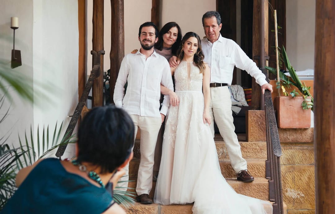 A woman in a white dress poses with three people on wooden steps while another person takes a photo. The setting appears to be indoors with plants in the foreground.