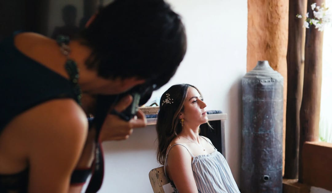 A woman sits on a chair, posing for a photographer who is focusing the camera on her in an indoor setting.