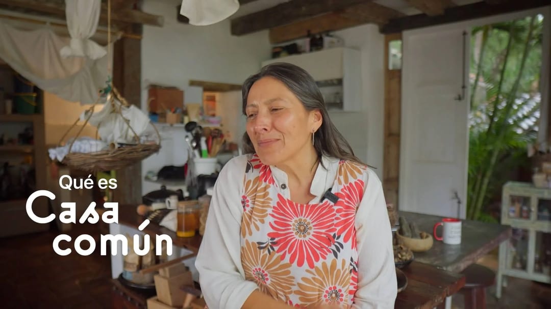 Una mujer con delantal floral sonríe en una cocina rústica de Casa Común.