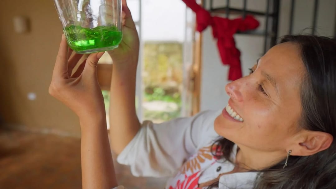 A smiling woman holds up a glass jar of bright green liquid to the light in a sunlit Casa Común doorway.