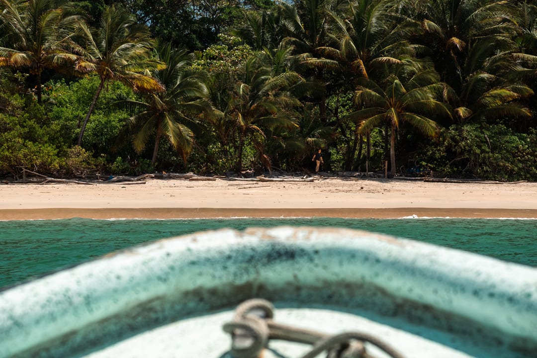 The prow of a small wooden boat approaching a remote palm-lined beach in Panama, with a single figure standing at the treeline.