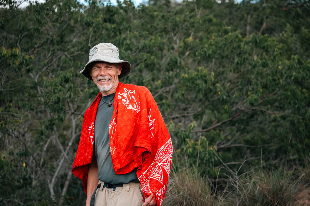 A smiling man in a hat wears a red patterned blanket in a wooded landscape.