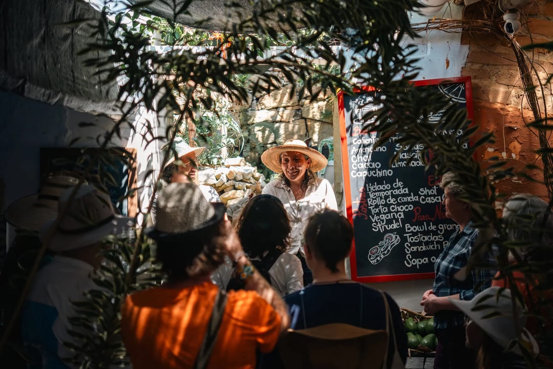 A woman in a hat explains something to a group gathered outside a local establishment.