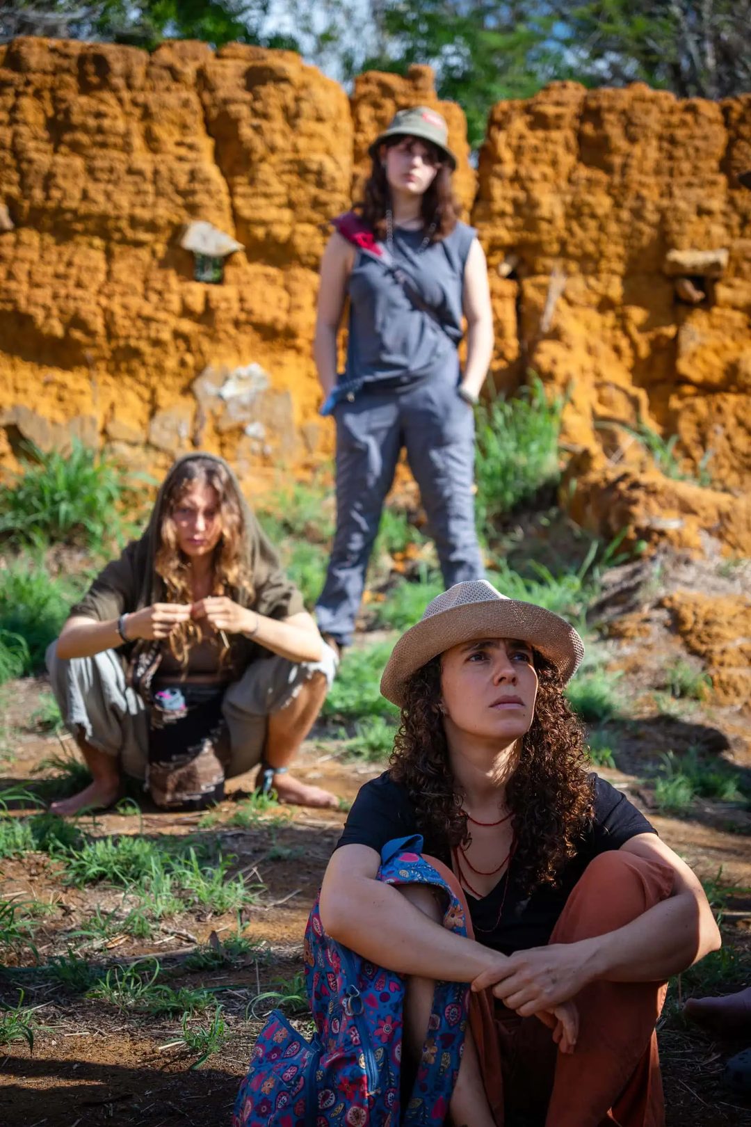 Three seated participants listen beside an earthen wall during a field activity.