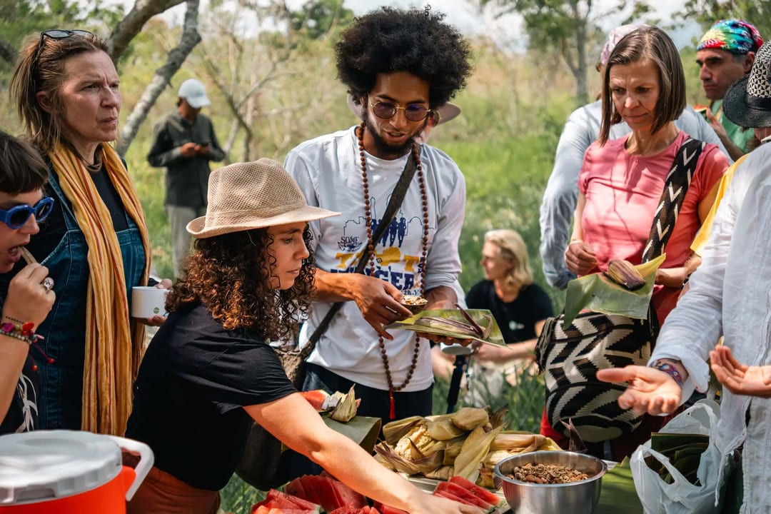 A group shares a communal meal with tamales around an outdoor table in a natural setting.