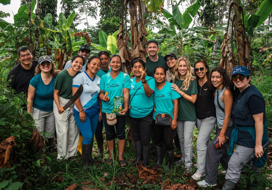 A large group of producers and visitors stand together for a group photograph among banana plants, one person holding up a packet of plantain chips.