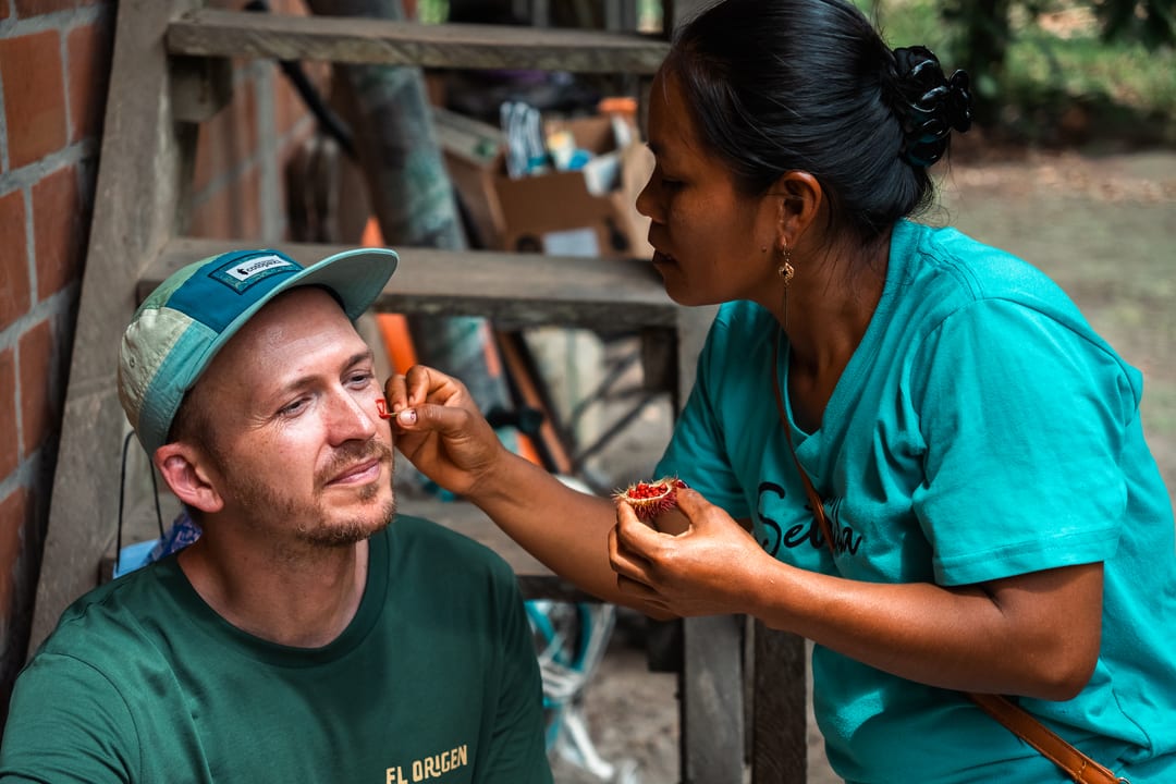 A woman in a turquoise shirt paints red markings onto the face of a seated man in an El Origen T-shirt, using pigment from a small bowl.