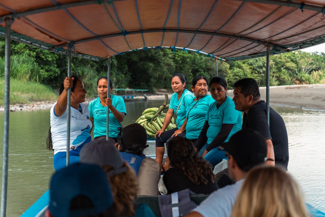 Passengers sit along both sides of a covered wooden riverboat, a bunch of green plantains stowed between them, as it moves along a river.