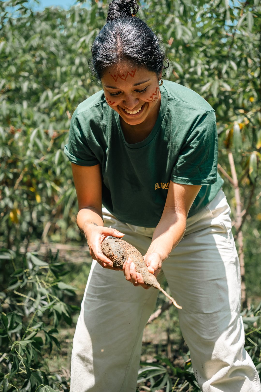 A smiling woman with red face paint bends forward in a sunlit field, holding a freshly dug root in both hands.