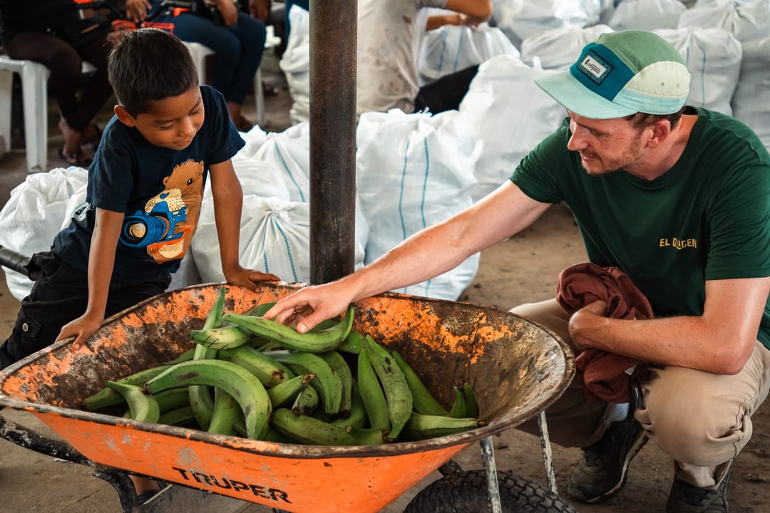 A young boy and a man in an El Origen T-shirt crouch either side of an orange wheelbarrow filled with green plantains, in a shed stacked with white sacks.