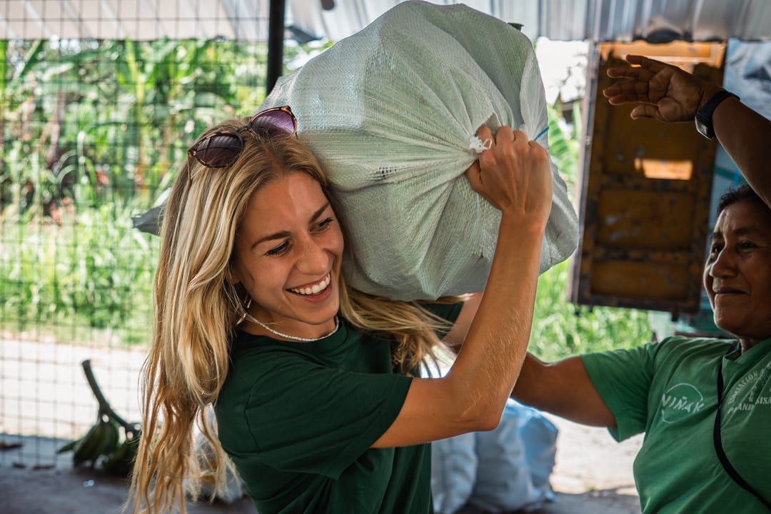 A laughing woman in a green T-shirt hoists a full white sack onto her shoulder while another woman steadies it from the right.