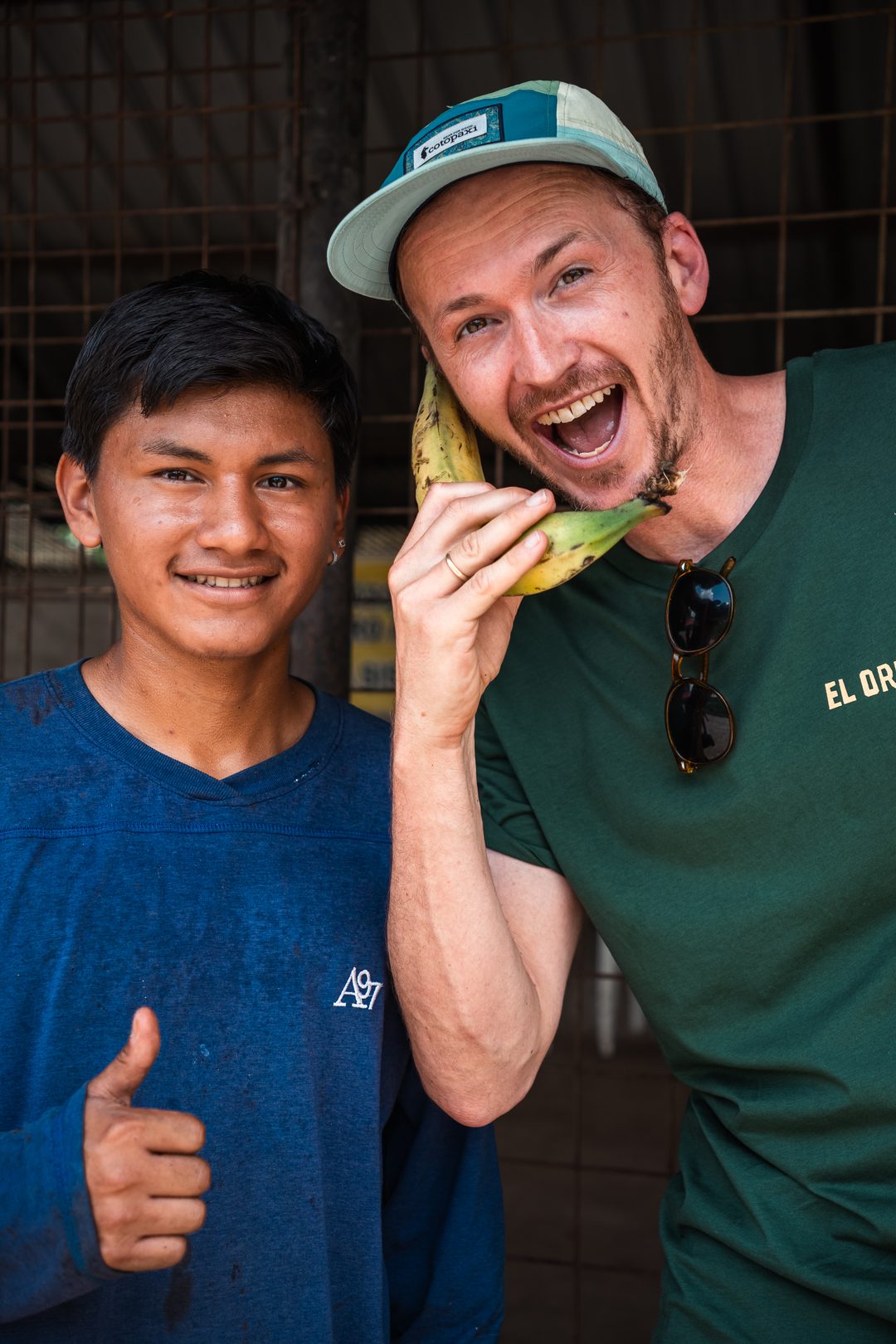 A laughing man in a cap holds a plantain to his ear like a telephone beside a young man in a blue shirt giving a thumbs up.