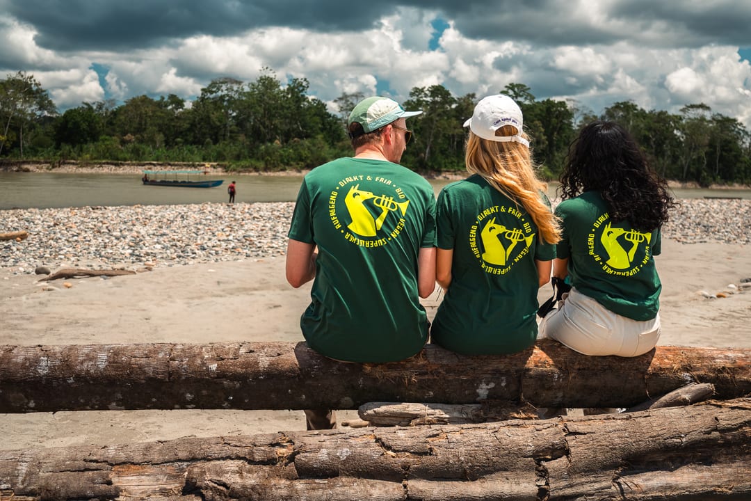 Three people in green El Origen T-shirts sit with their backs to the camera on a fallen log, looking out over a wide stony riverbank under a cloudy sky.