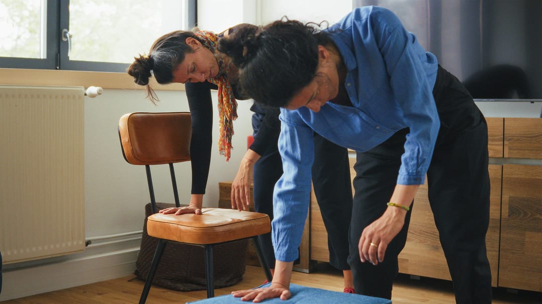 Two women perform a stretching exercise indoors, each leaning forward with one hand on a supportive surface and the other arm extended downward.