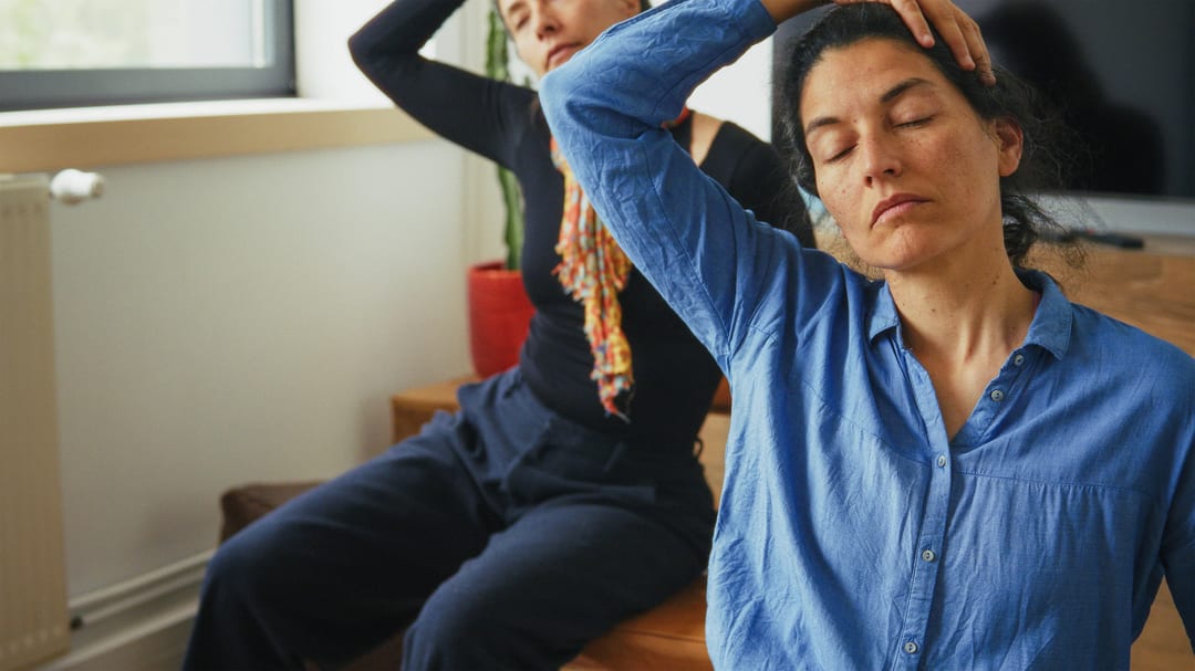 Two people seated indoors with eyes closed, gently stretching their necks by tilting their heads to one side and using a hand to assist the stretch.