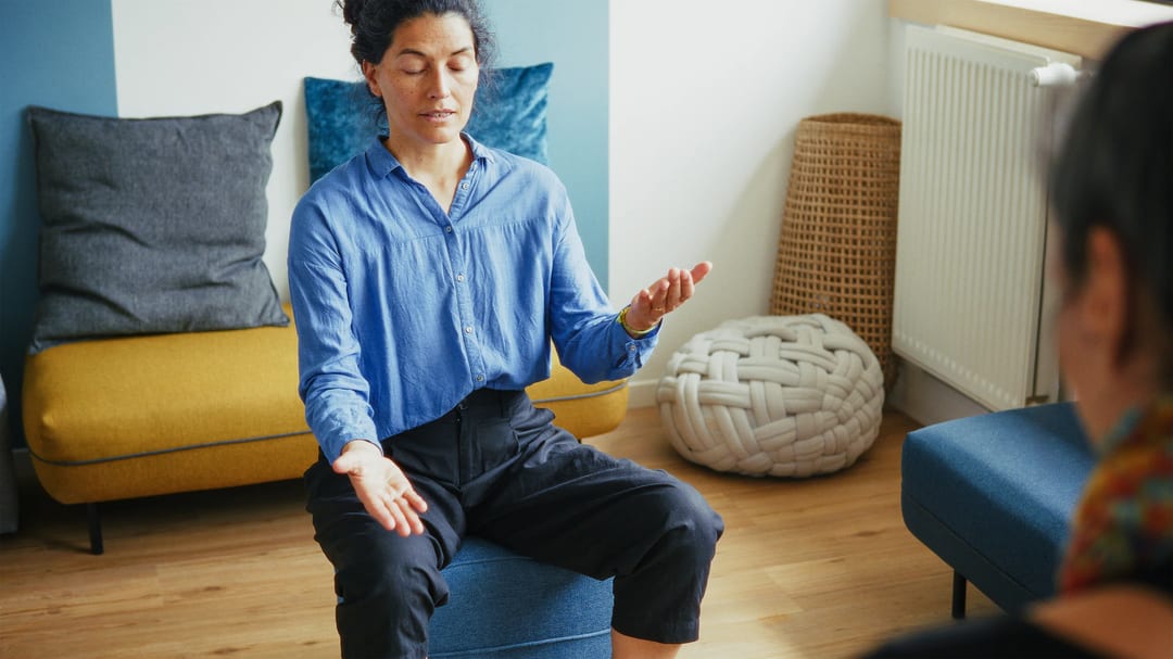 A person sits on a blue chair with eyes closed and arms open, appearing to meditate or practice mindfulness in a cozy, modern room.