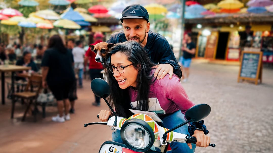 Cris en una moto colorida con Marty y su perro en un mercado al aire libre.