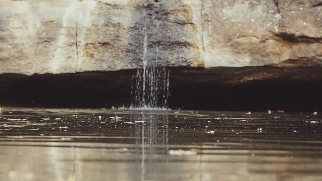 A small stream of water gently falls from a rocky ledge above into a calm, shallow pool below, creating ripples on the reflective water surface—a tranquil moment captured by Embodic Films.