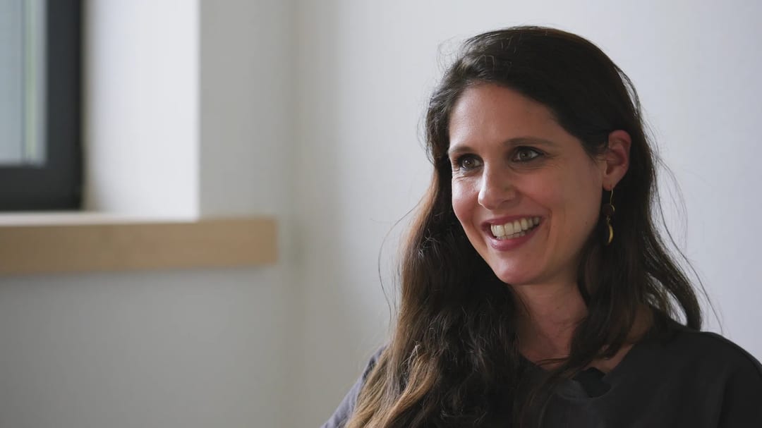 Smiling woman with long brown hair and gold earrings in a bright room.