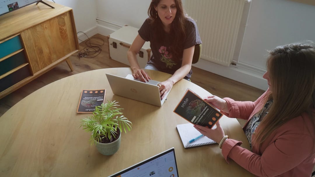 Two women collaborating at a table with Sindup materials and laptops.