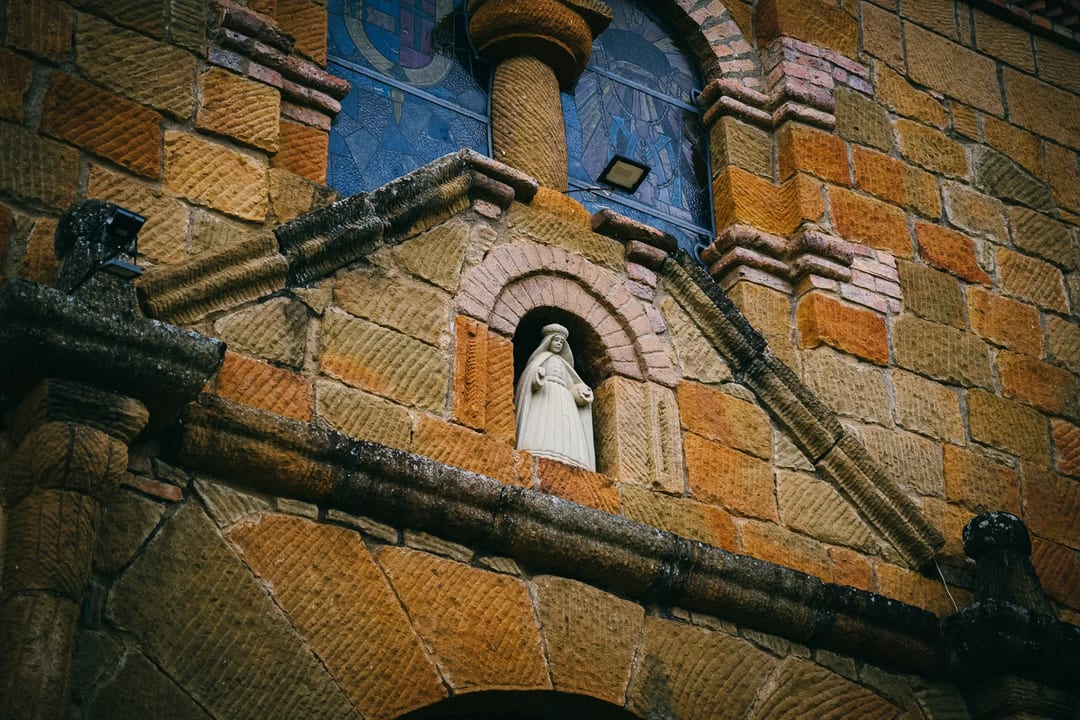 Stone church facade with Virgin Mary statue and stained glass window.