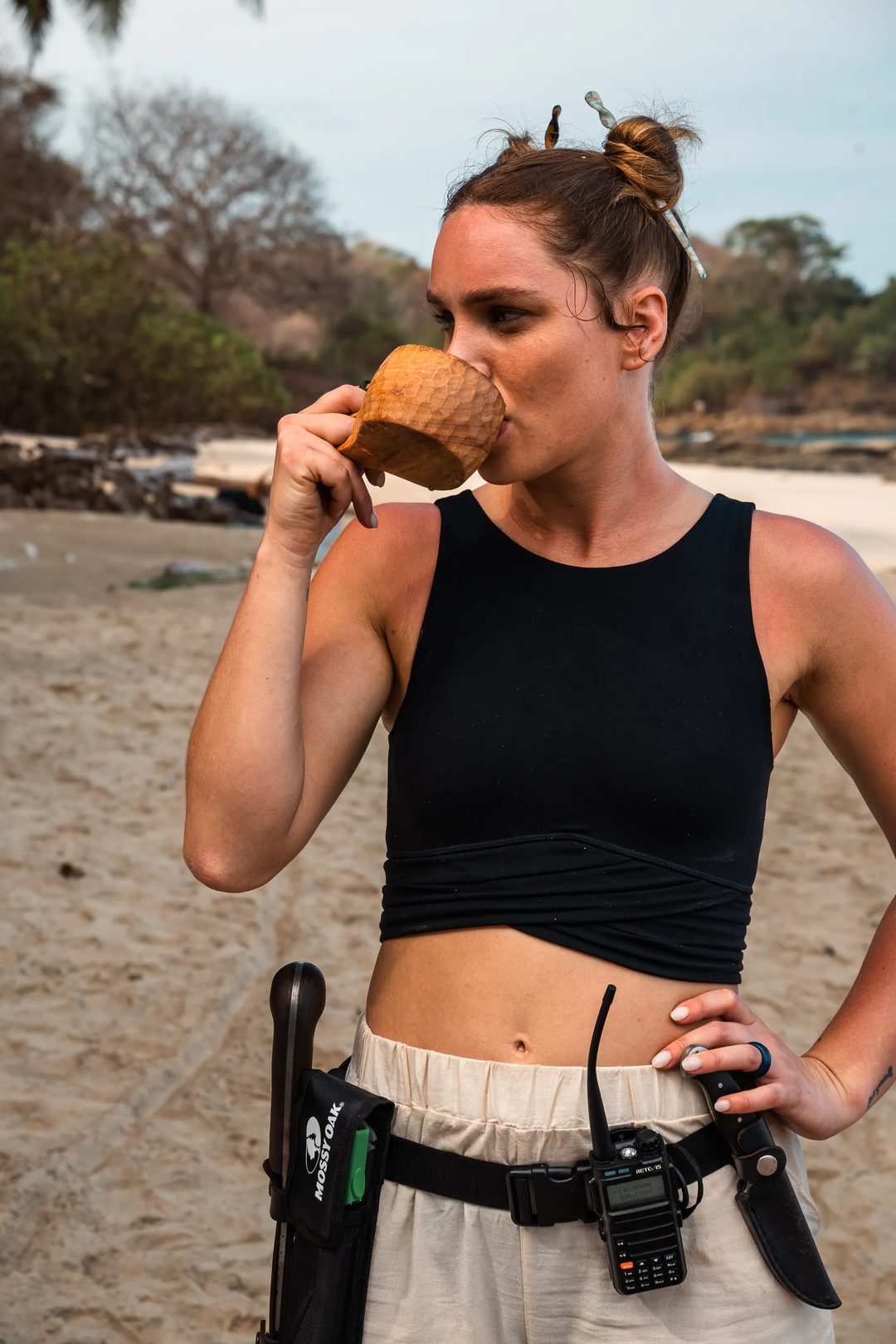 A woman in a black crop top and beige pants drinks from a coconut on a sandy beach, with a machete and walkie-talkie attached to her belt—capturing the adventurous spirit of Embodic Films.