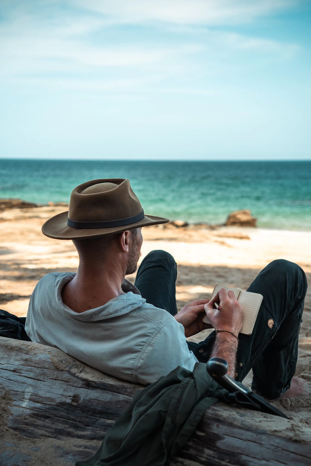 A person wearing a hat sits on a driftwood log at the beach, writing in a notebook while facing the ocean—a tranquil moment captured in true Embodic Films style.