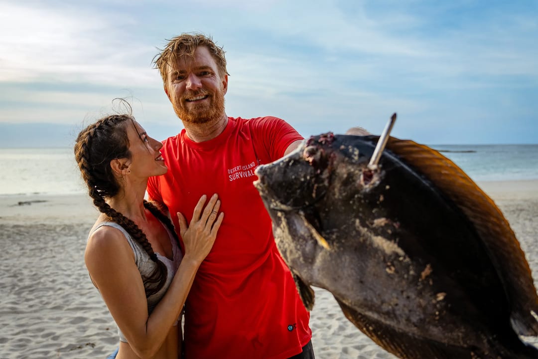 Couple on a beach displaying a large fish caught during their Desert Island Survival adventure.