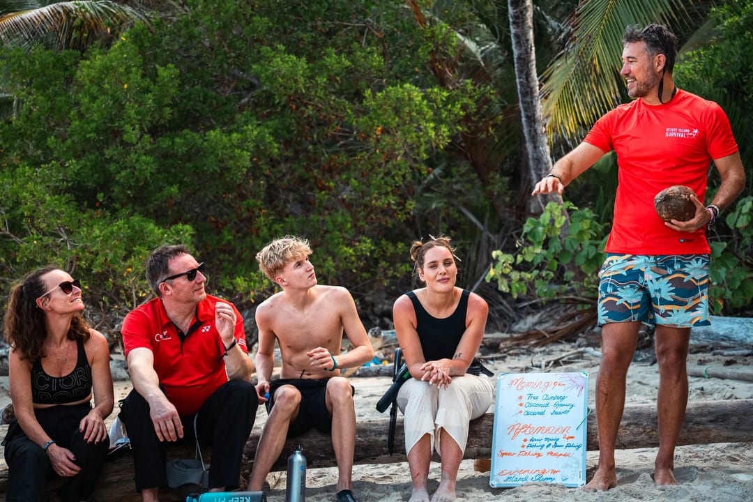 Five people sit on logs on a sandy beach; one person stands holding a coconut and appears to be speaking. A whiteboard with writing is visible in the background, capturing a behind-the-scenes moment from Embodic Films.