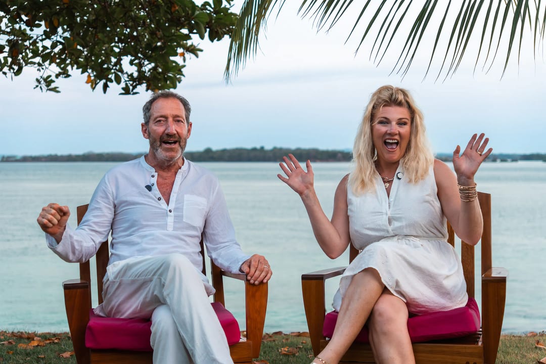 A man and a woman sit on wooden chairs by the water, both smiling and gesturing enthusiastically beneath palm leaves—a class=