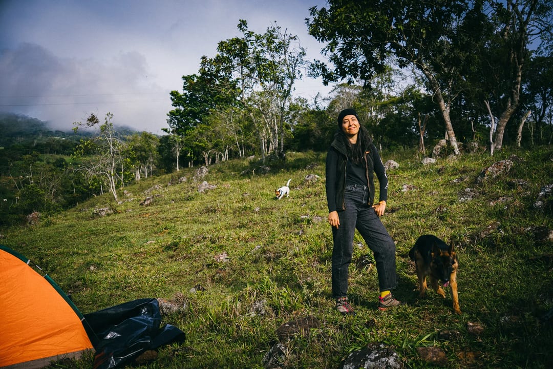 Woman camping in El Páramo with dogs, wearing a black jacket and jeans. Tent visible. Cloudy sky, green landscape.