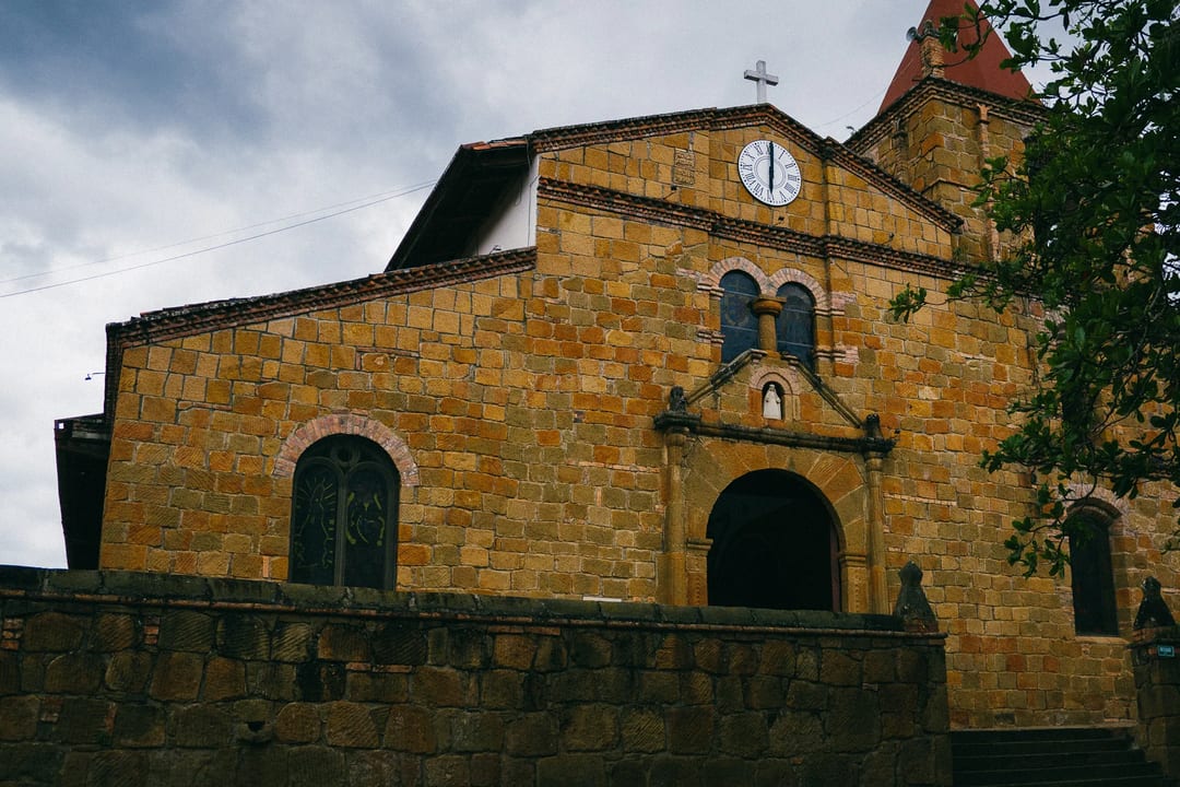 Facade of a yellow stone church with a clock and arched doorway under a cloudy sky.